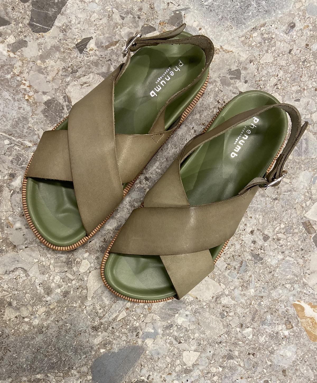 Olivengrønne sandaler fra phenumb copenhagen med krydsede stropper og justerbar rem, placeret på en stengulvbaggrund.