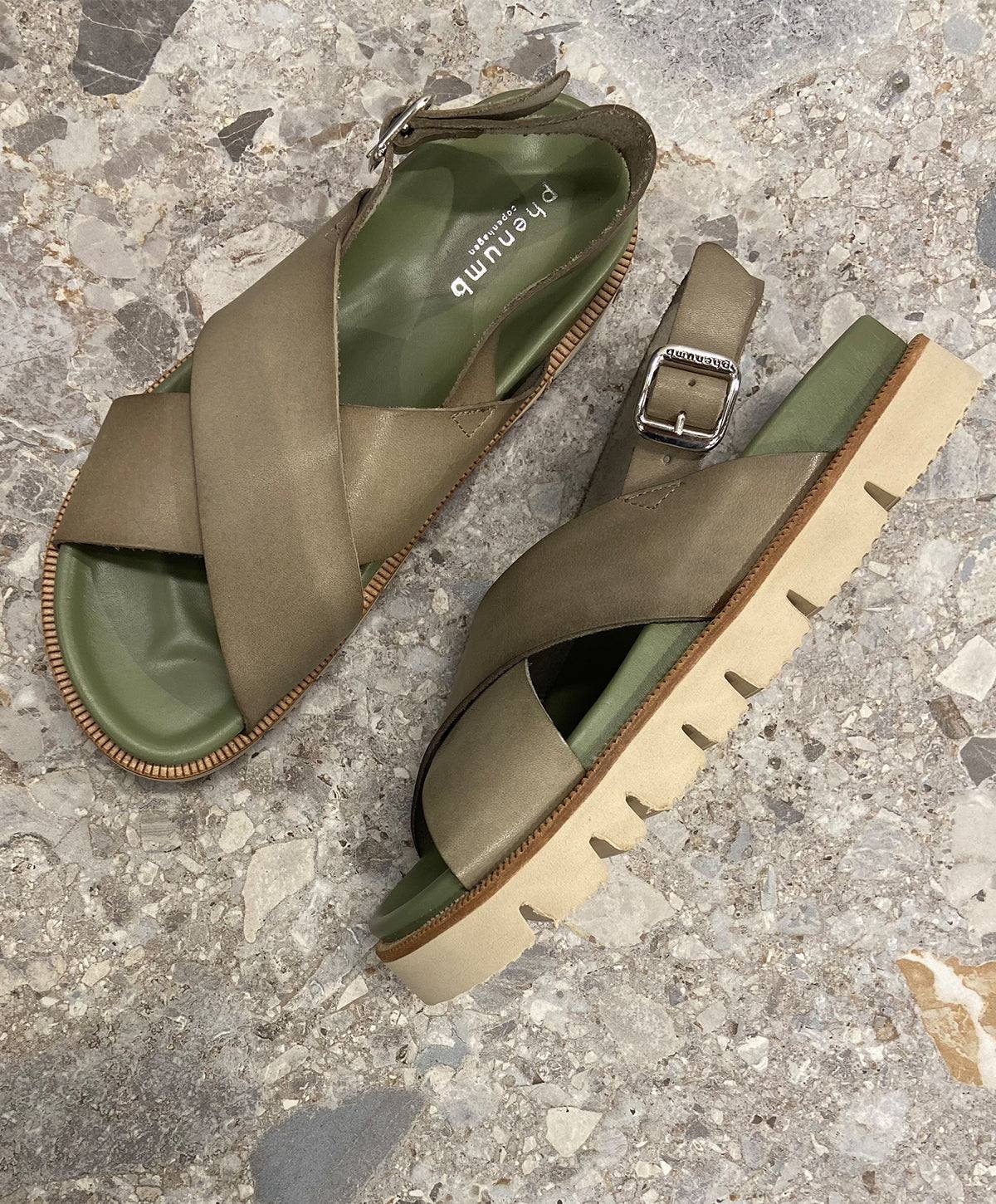 Grønne sandaler fra phenumb copenhagen med krydsede stropper og justerbar spænde, placeret på en stengulvbaggrund.